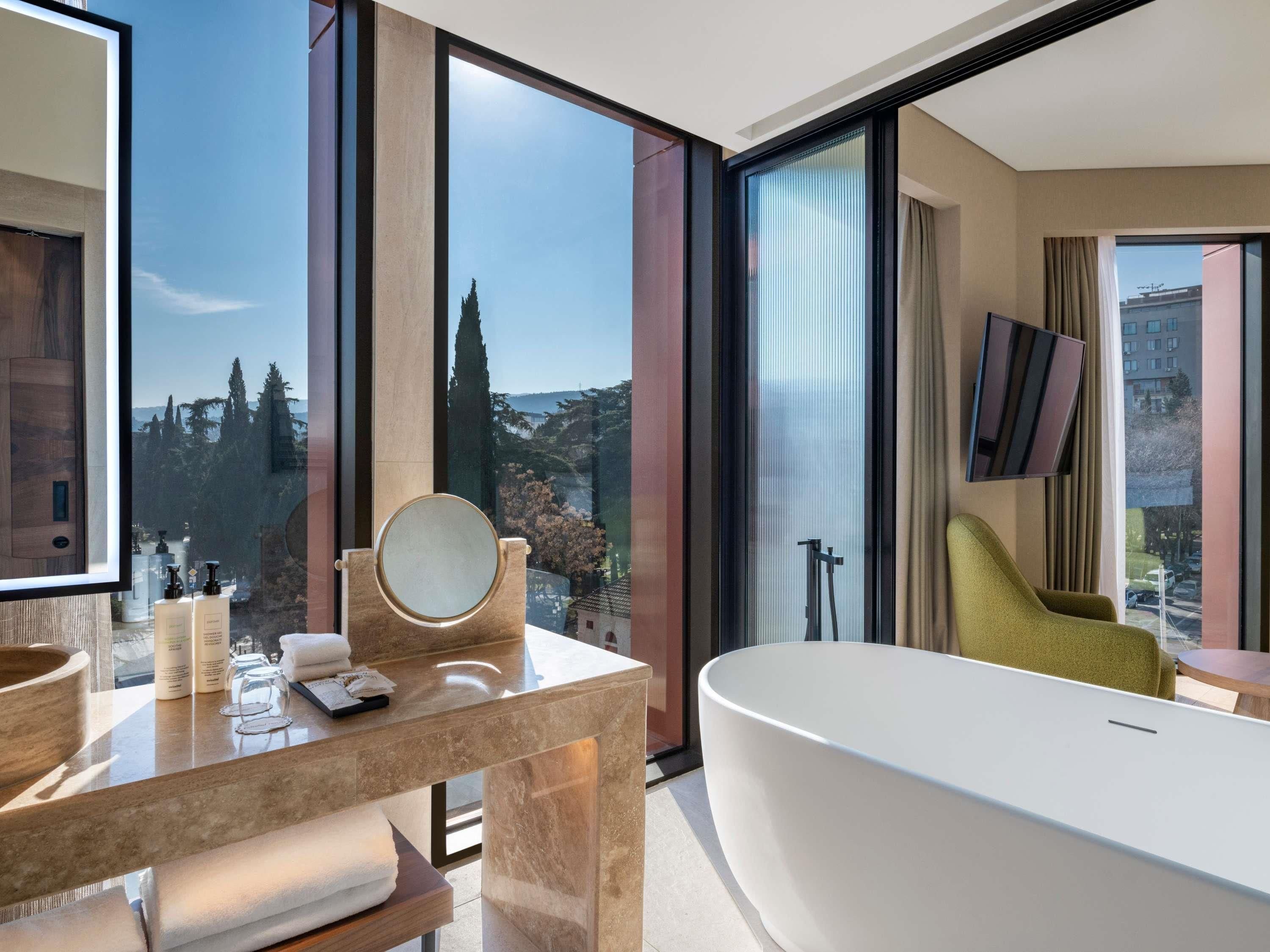 A modern bathroom with a large bathtub, a vanity with a mirror, and a view of the city through large windows.