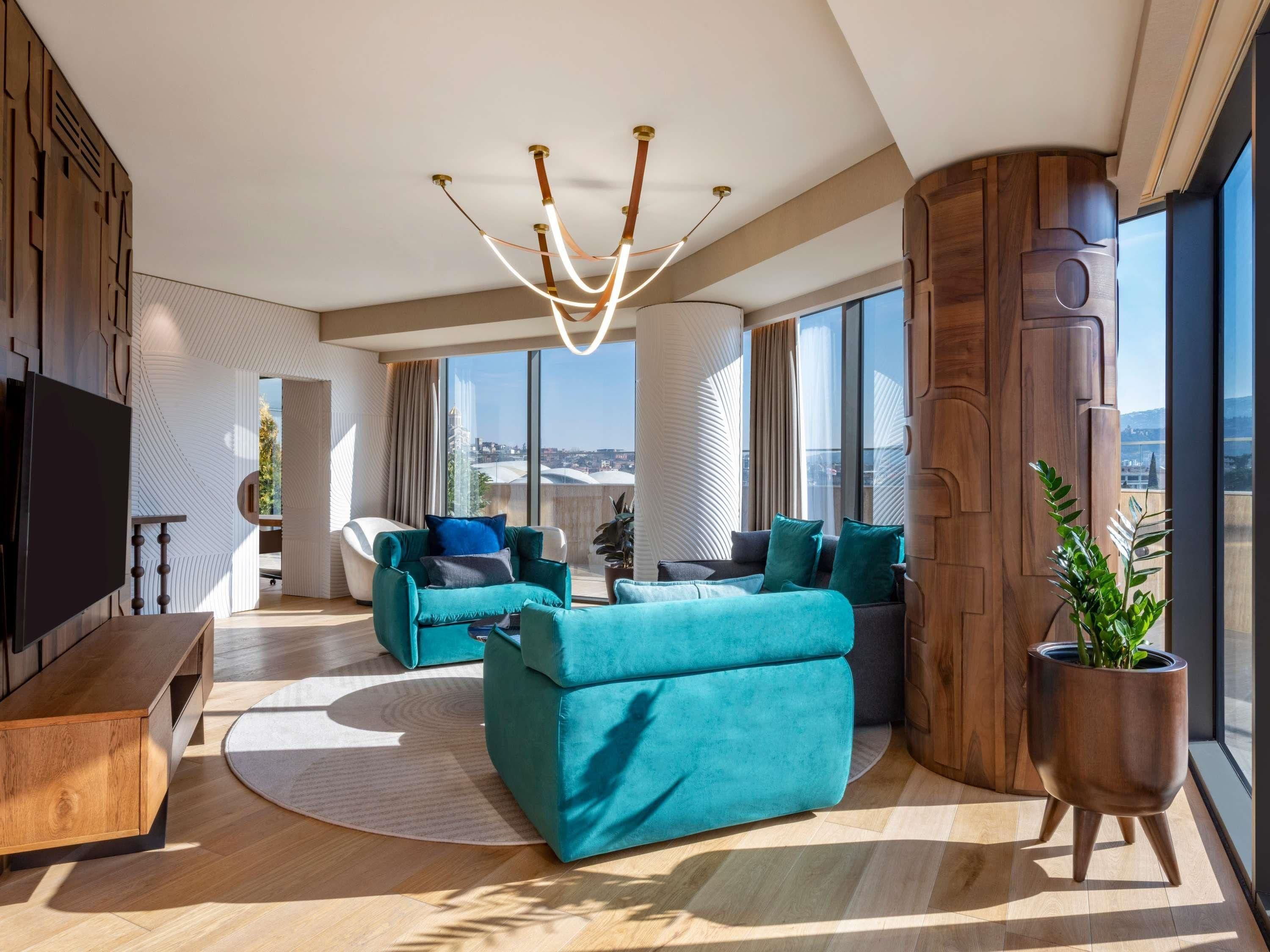 A modern living room with a large TV, teal sofas, a unique ceiling light, and a large window with a city view.