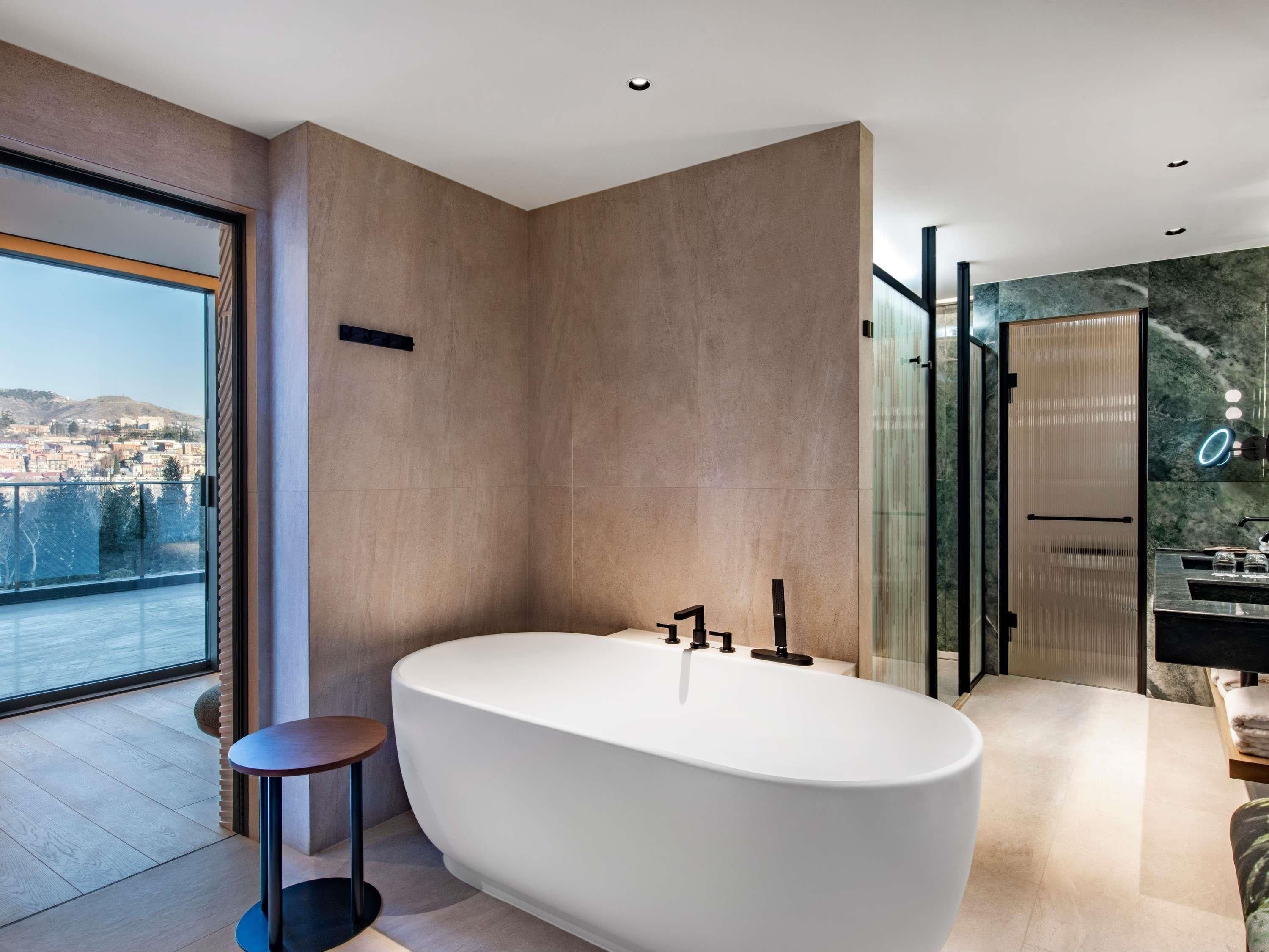A modern bathroom with a freestanding bathtub, a glass shower enclosure, and a view of a cityscape through a large window.