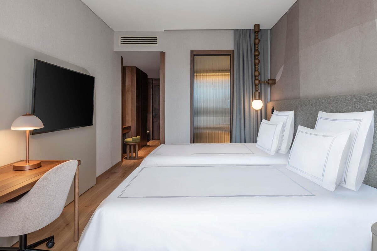 A hotel room with a bed, a desk with a chair, a TV mounted on the wall, and a window with curtains.