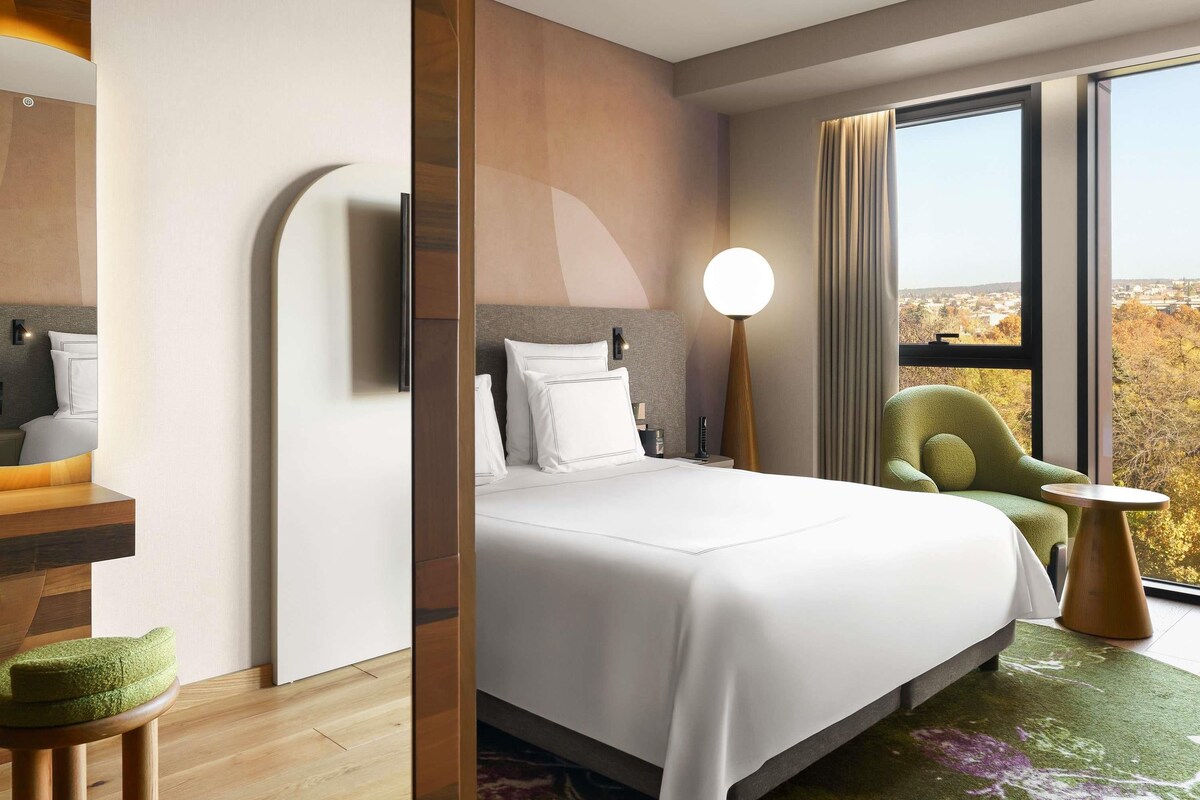A modern hotel room with a bed, a green chair, a round table, and a view of the city.