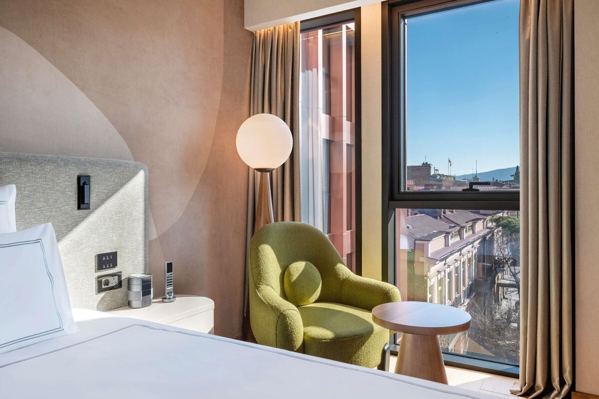 A hotel room with a bed, a green armchair, a small round table, a lamp, and a large window with a city view.