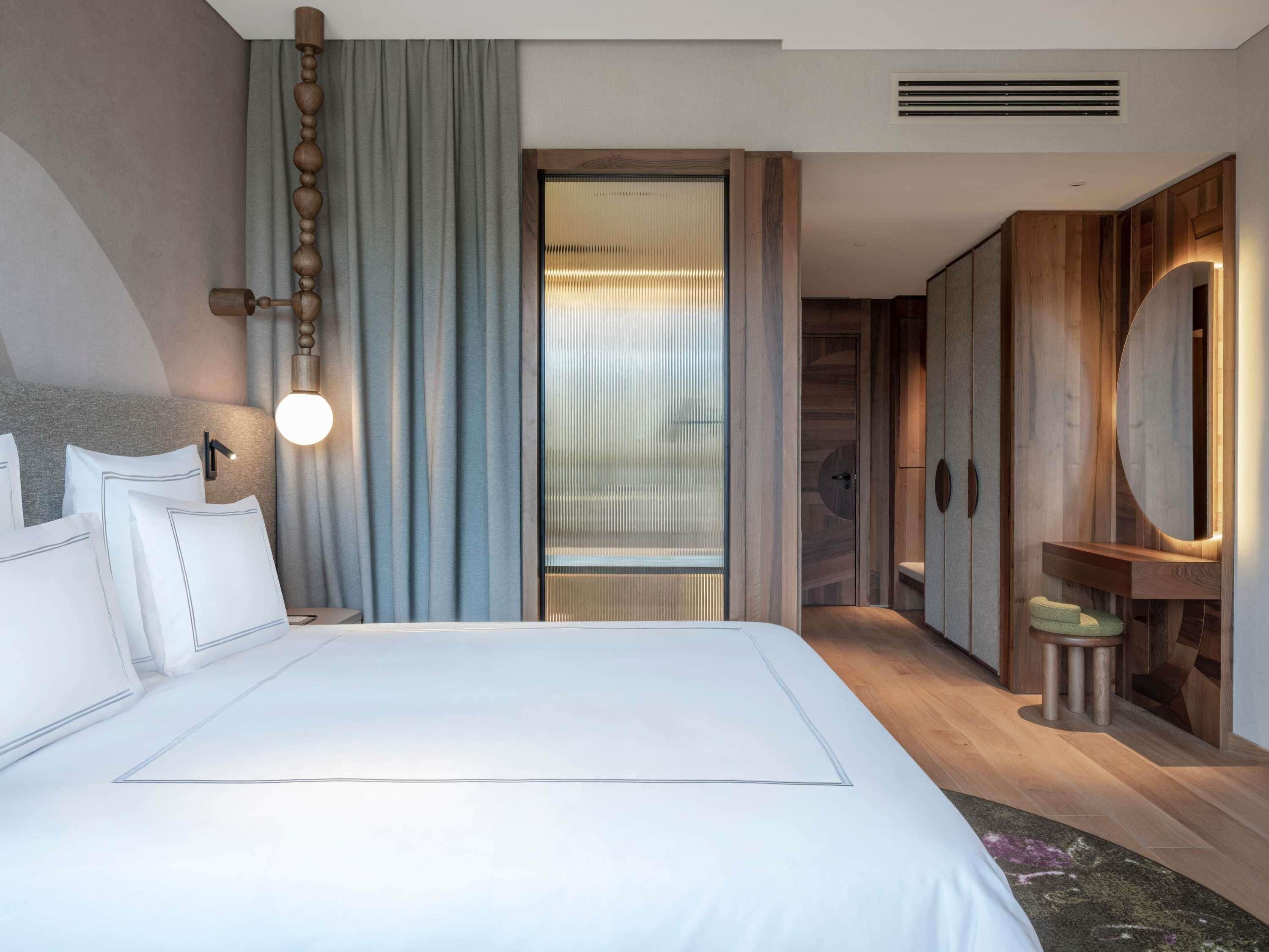 Een moderne hotelkamer met een groot bed, een wandlamp, een houten garderobe en een schuifdeur naar een badkamer.