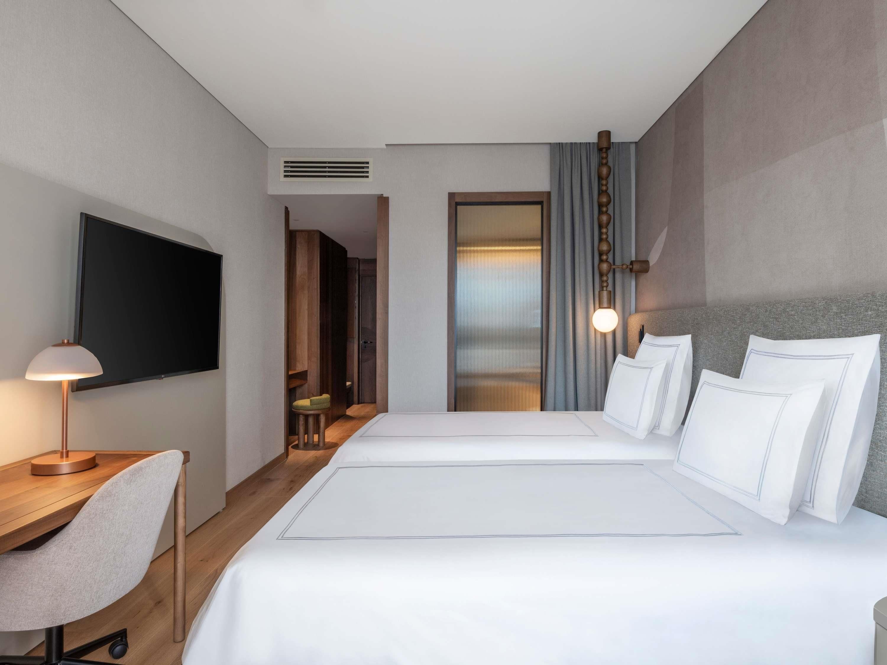 Een hotelkamer met een bed, een bureau met stoel, een aan de muur bevestigde televisie en een raam met gordijnen.
