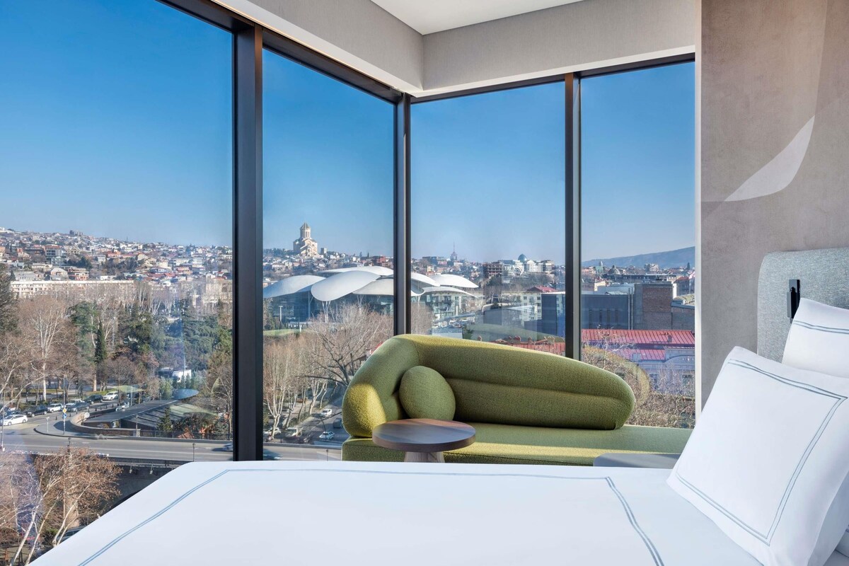 A modern hotel room with a large window offering a city view, a green sofa, and a bed with white bedding.