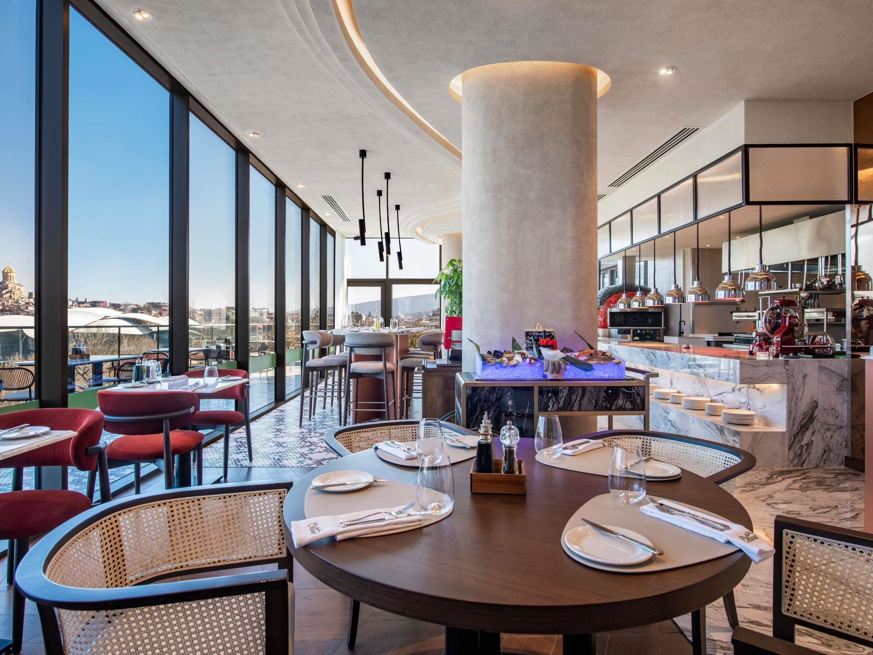 En modern restaurang med runda bord, röda stolar och utsikt över staden.