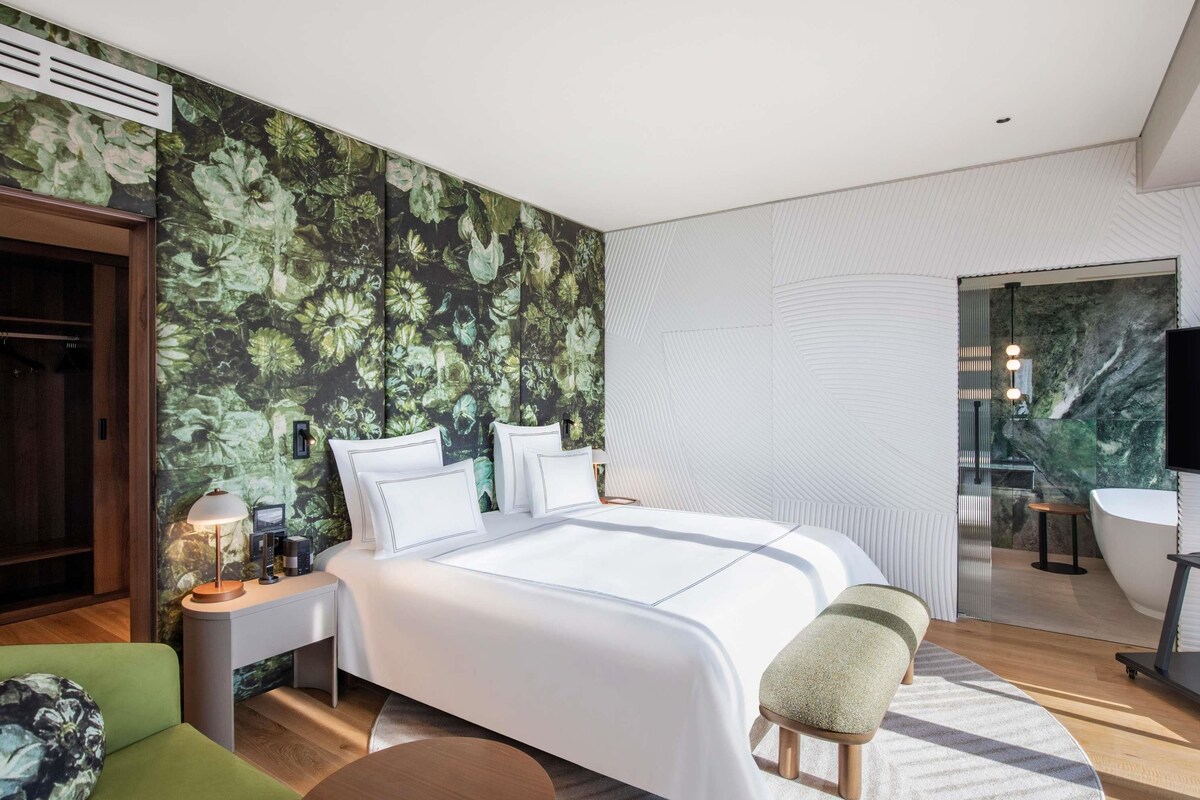 Een moderne slaapkamer met een groot bed, een bureau, een groene stoel en een badkamer zichtbaar door een geopende deur.