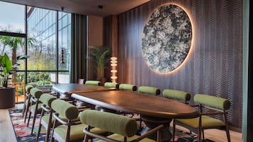 Una sala de reuniones moderna con una gran mesa de madera, sillas tapizadas en verde, un muro decorativo con estampado floral y una planta en maceta.