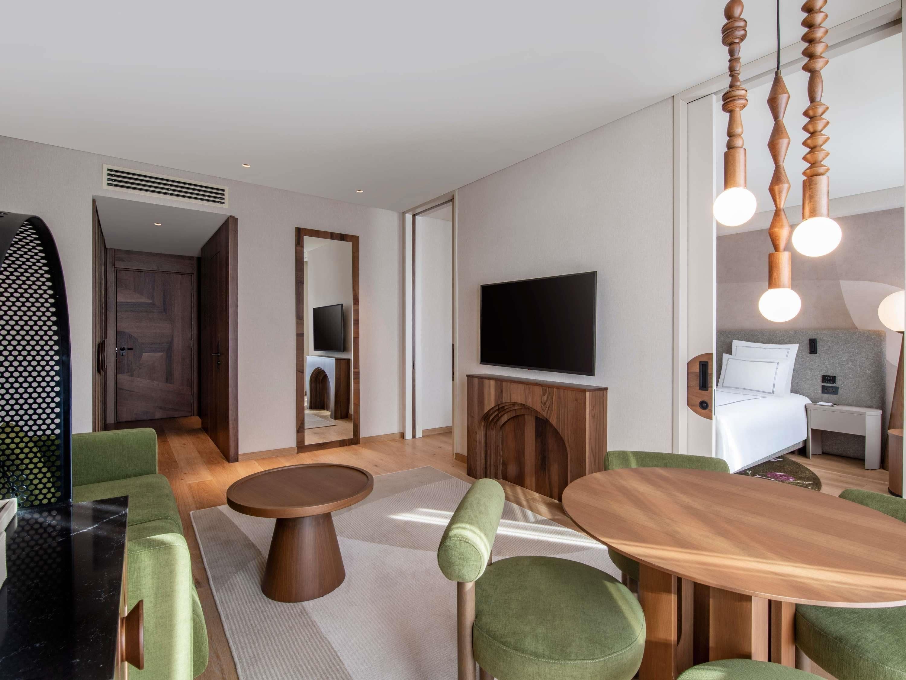 A modern hotel room with a wooden dining table, green upholstered chairs, a flat-screen TV, and a bed with a grey headboard.
