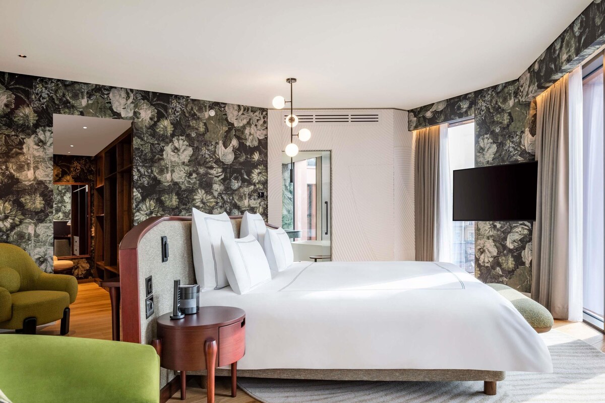 A modern hotel room with a large bed, a green armchair, a small wooden side table, and a flat-screen TV mounted on the wall.