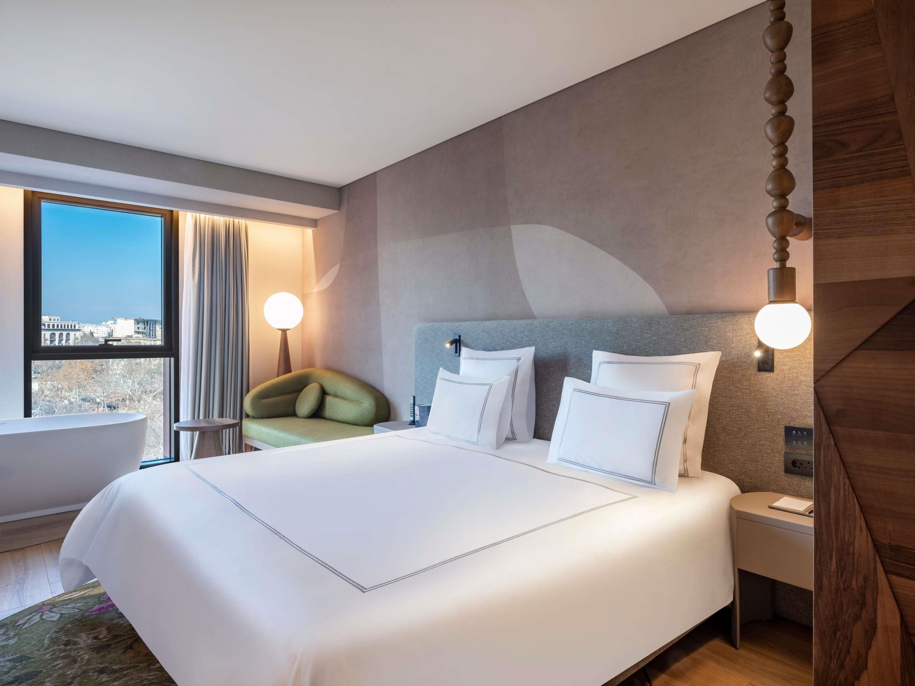 A hotel room with a large bed, a green chair, a nightstand, and a view of the city through a large window.