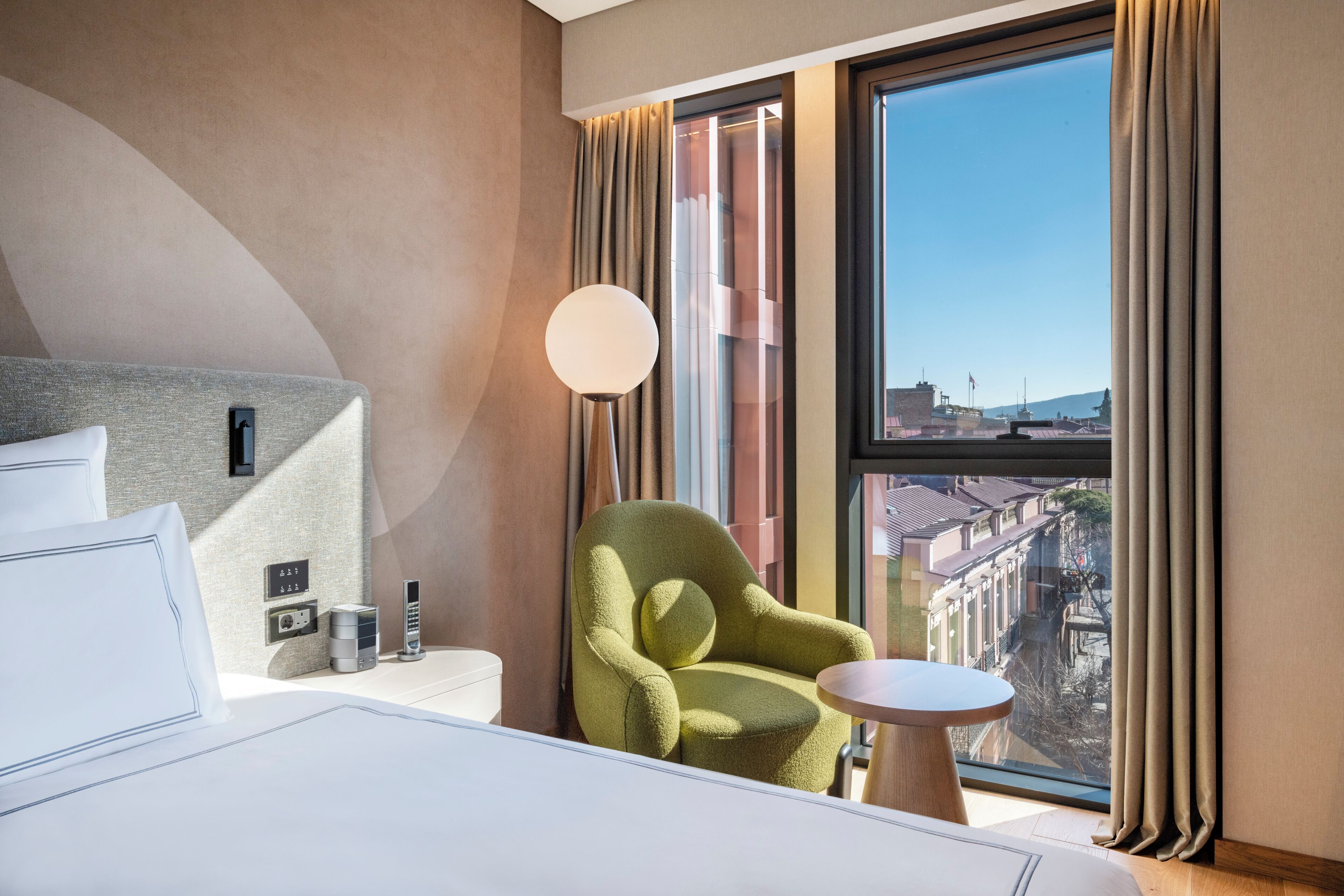 A hotel room with a bed, a green armchair, a small table, a lamp, and a large window with curtains.