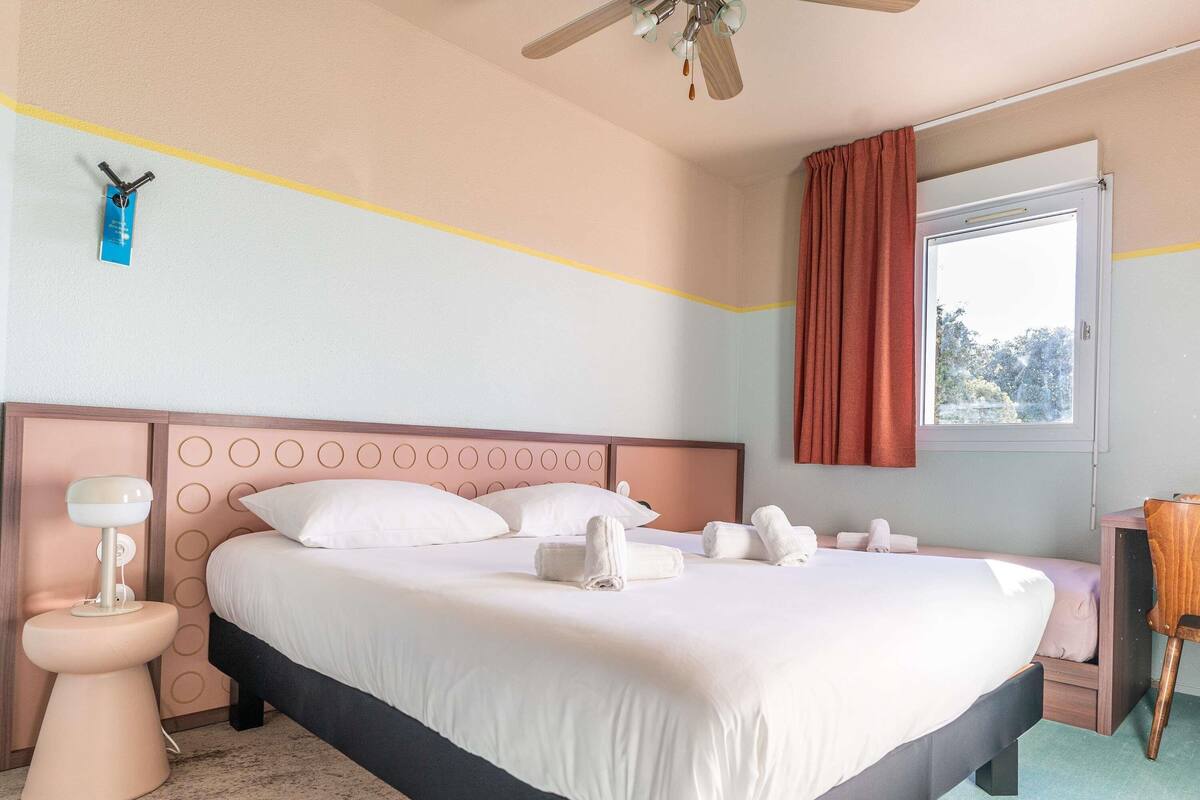Ein Hotelzimmer mit Bett, Nachttisch, Lampe und einem Fenster mit Vorhängen.