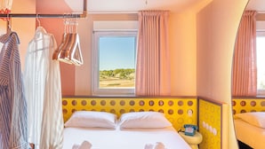 Une chambre d’hôtel avec un lit, deux oreillers, une fenêtre avec des rideaux, un portant à vêtements et une table de chevet avec un téléphone.