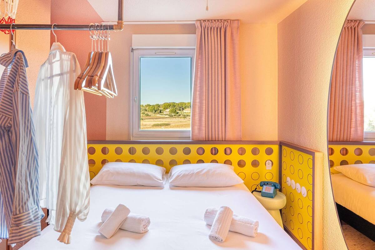 Ein Hotelzimmer mit Bett, zwei Kissen, einem Fenster mit Vorhängen, einem Kleiderhaken und einem Nachttisch mit Telefon.