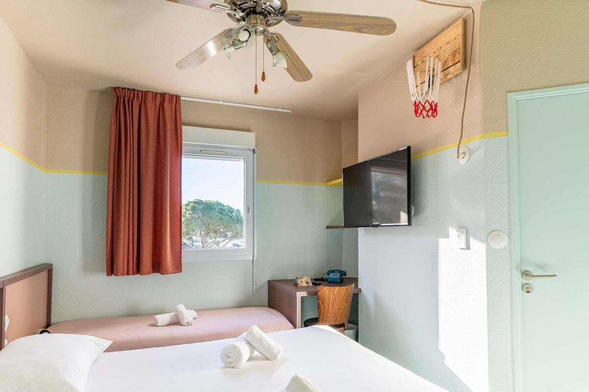 Ein Hotelzimmer mit Bett, Deckenventilator, an der Wand montiertem Fernseher, einem Fenster mit Vorhängen und einer Tür.