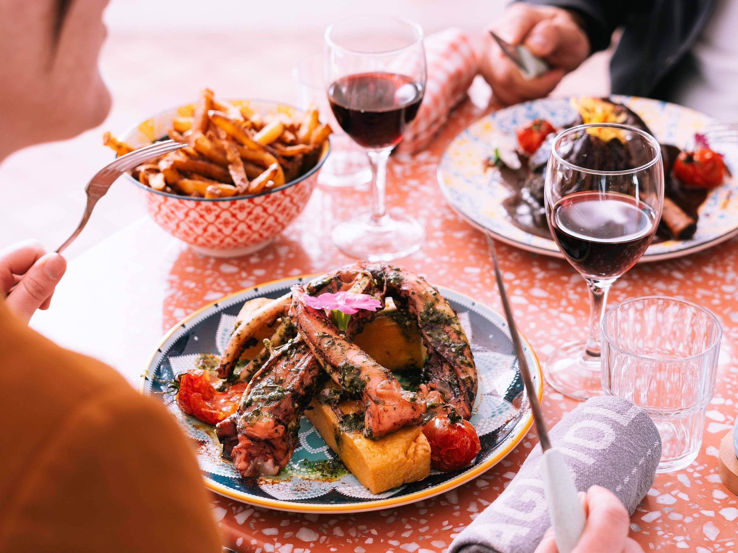 Una mesa de comedor con platos de comida, copas de vino tinto y un bol de papas fritas.