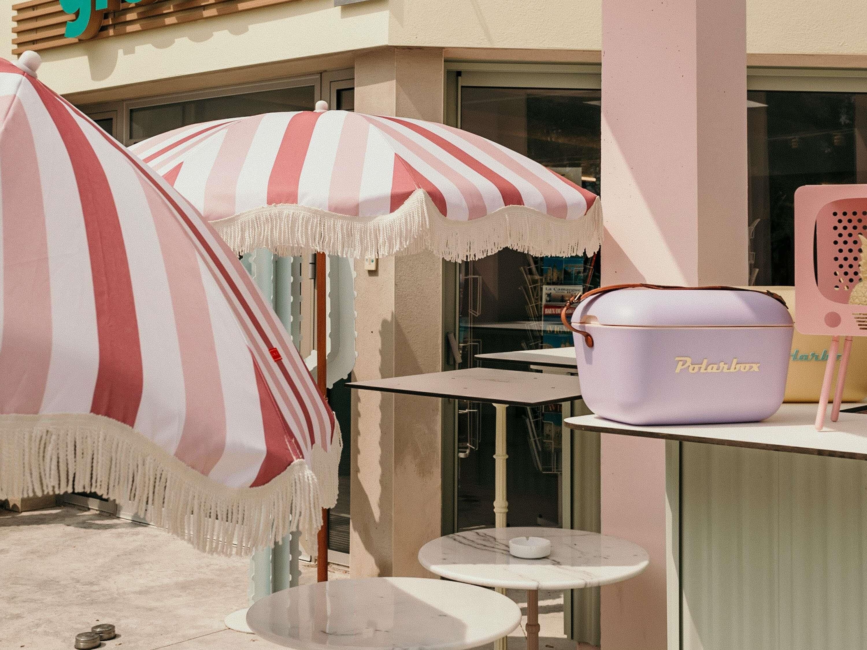 Una pequeña zona de estar al aire libre con sombrillas a rayas, una mesa con superficie blanca y una bolsa morada con la etiqueta "Polarbox" sobre un soporte.