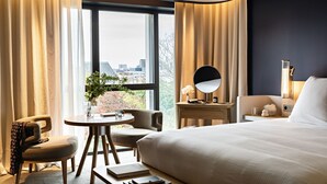 A hotel room with a large bed, a desk with a chair, a round table with a plant, and a view of the city through large windows. - Domaine de la Reine Margot Paris-Issy - MGallery Collection (Issy-les-Moulineaux)