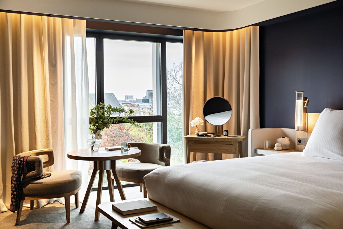 A hotel room with a large bed, a desk with a chair, a round table with a plant, and a view of the city through large windows.