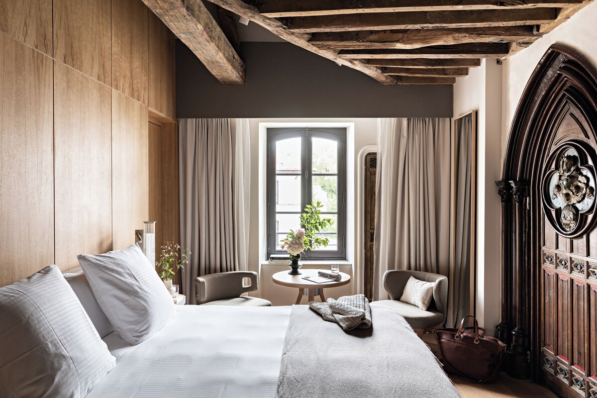 A hotel room with a large bed, a small table with a vase and a glass, two chairs, and a wooden ceiling.