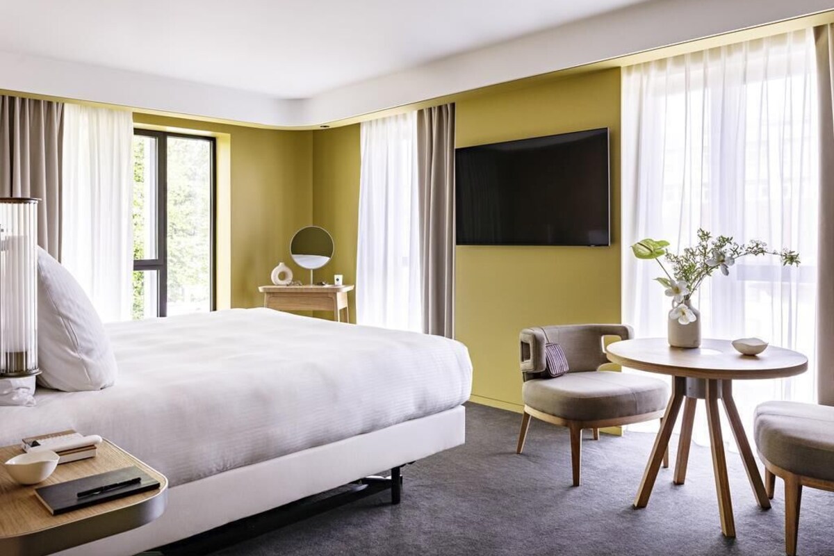 A hotel room with a bed, bedside table, lamp, chair, round table, and a TV.