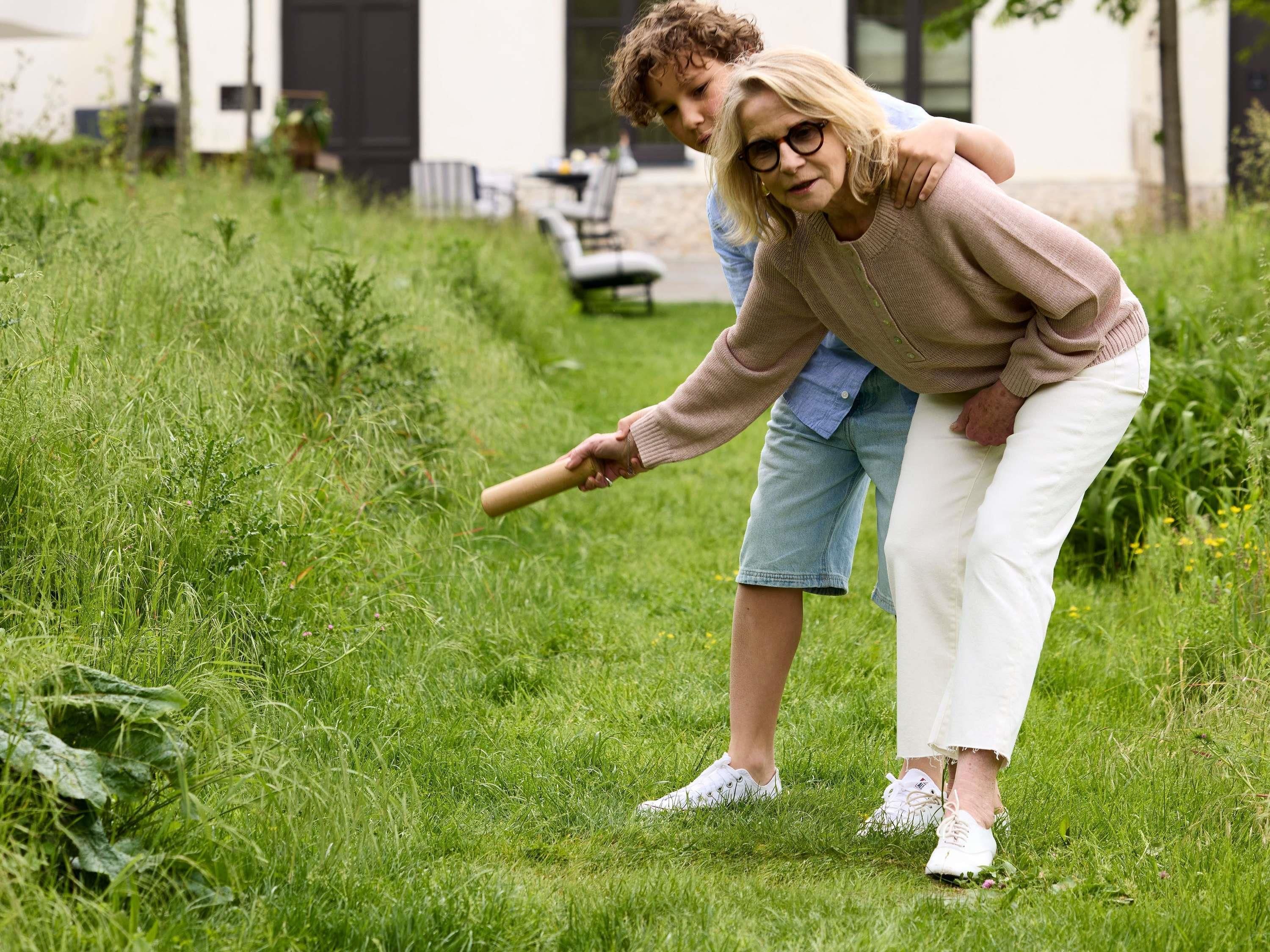 A person in a beige sweater and white pants holding a stick, standing in a grassy area with a building in the background.