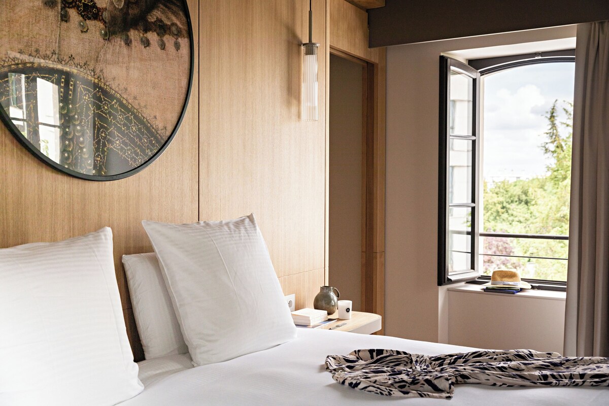 A hotel room with a bed, pillows, a bedside table with a lamp, a window with a view, and a decorative mirror.