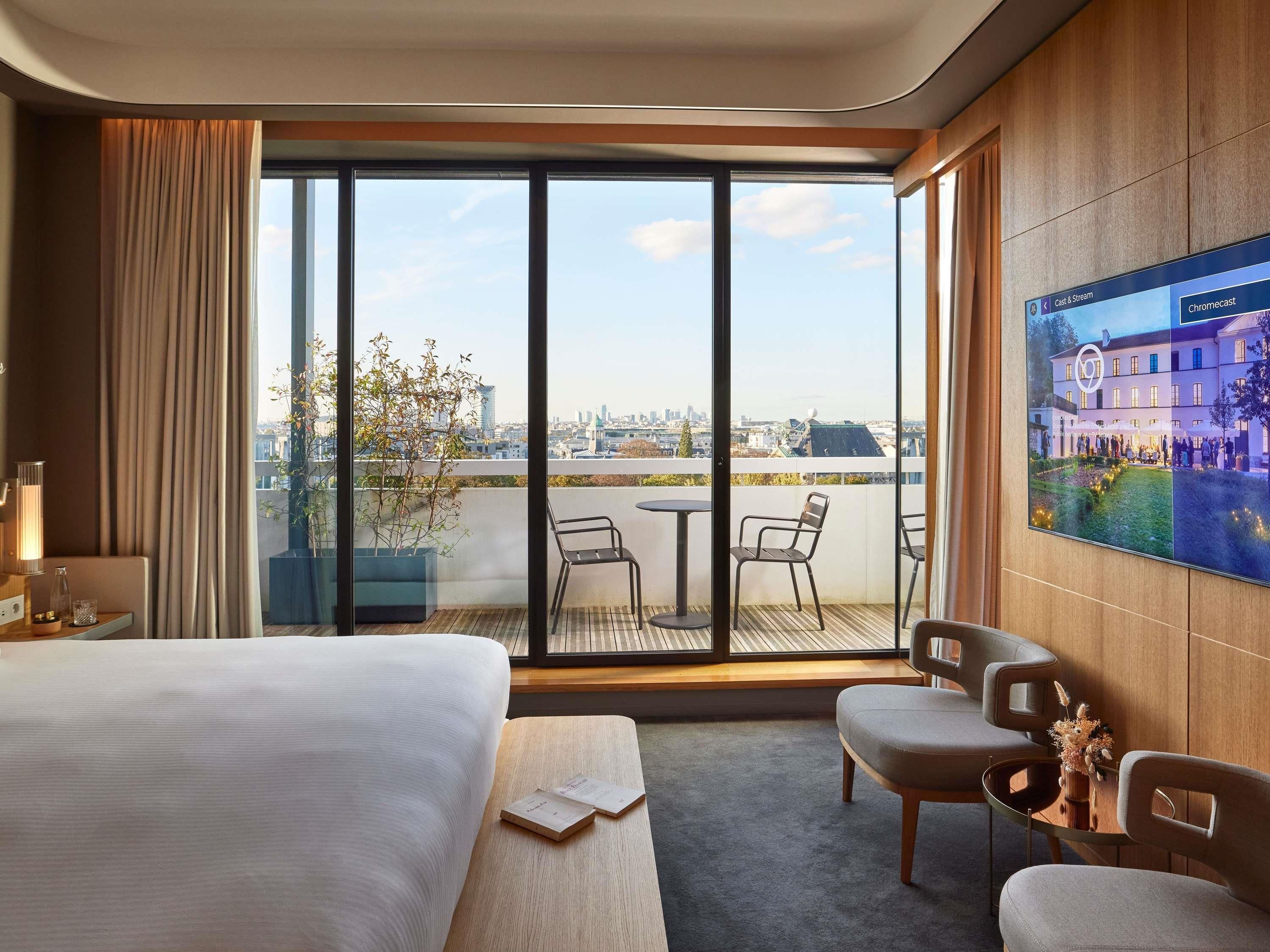 A hotel room with a large bed, a small table, two chairs, a potted plant, and a large window offering a city view.