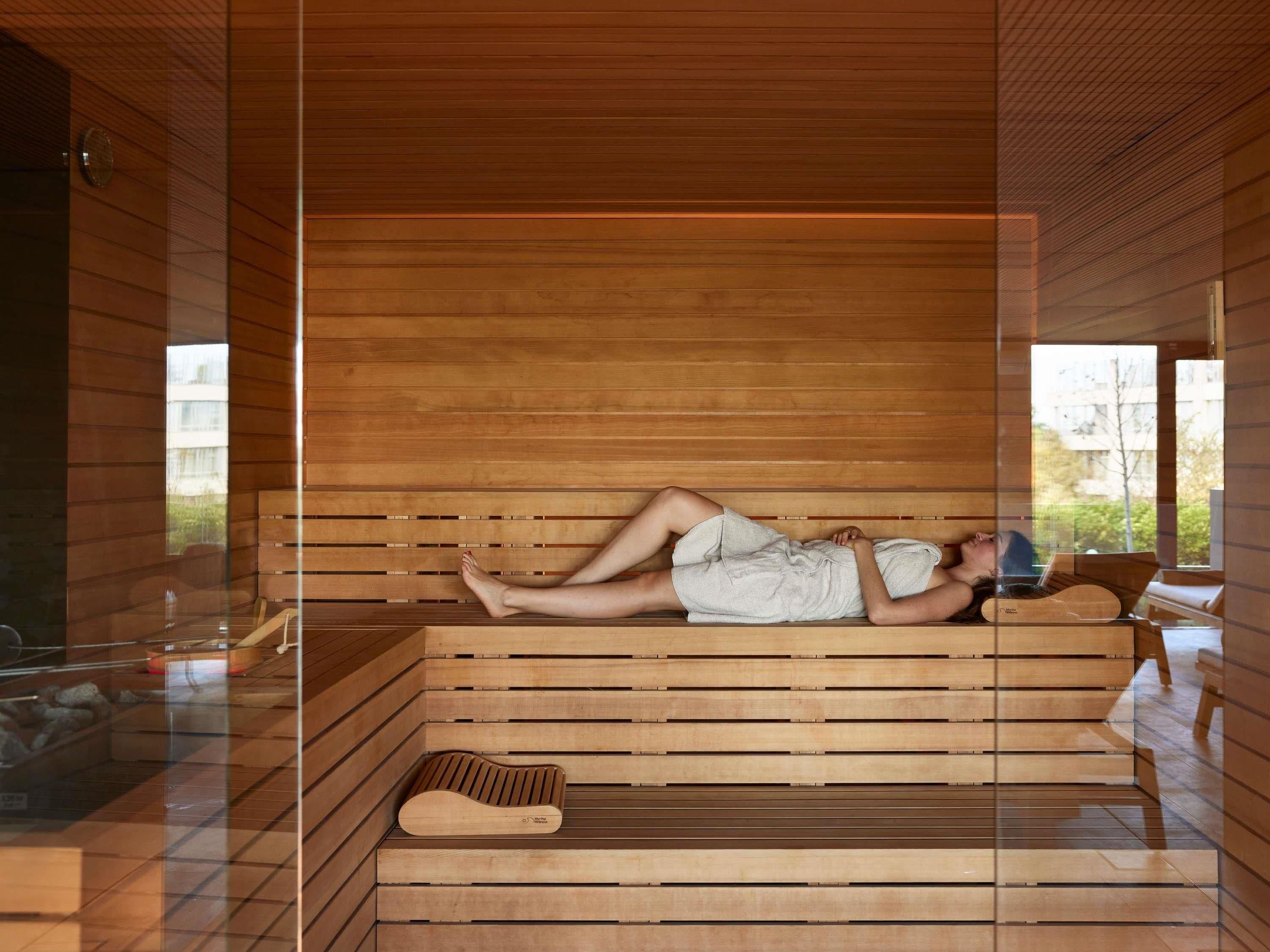 Iemand ligt op een houten bank in een kamer met een glazen wand, houten latten in het plafond en een bank met kussen.