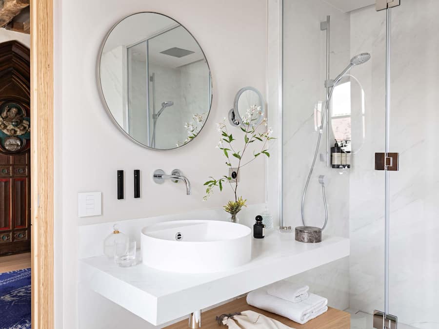 A modern bathroom with a round sink, a large mirror, a shower, and a vase with flowers.