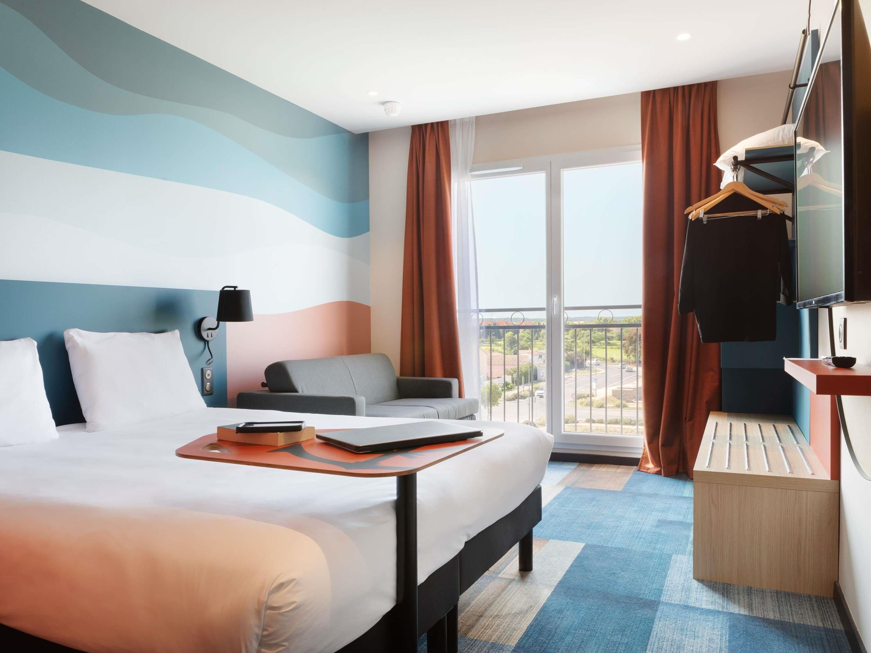 Habitación de hotel con una cama grande, un sofá gris, un televisor y un balcón con vistas al campo.