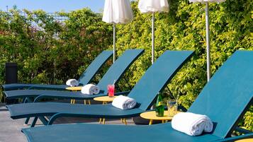 A row of blue lounge chairs with white towels, a glass of pink drink, and a bottle on a small round table, set against a backdrop of green foliage and a clear sky.
