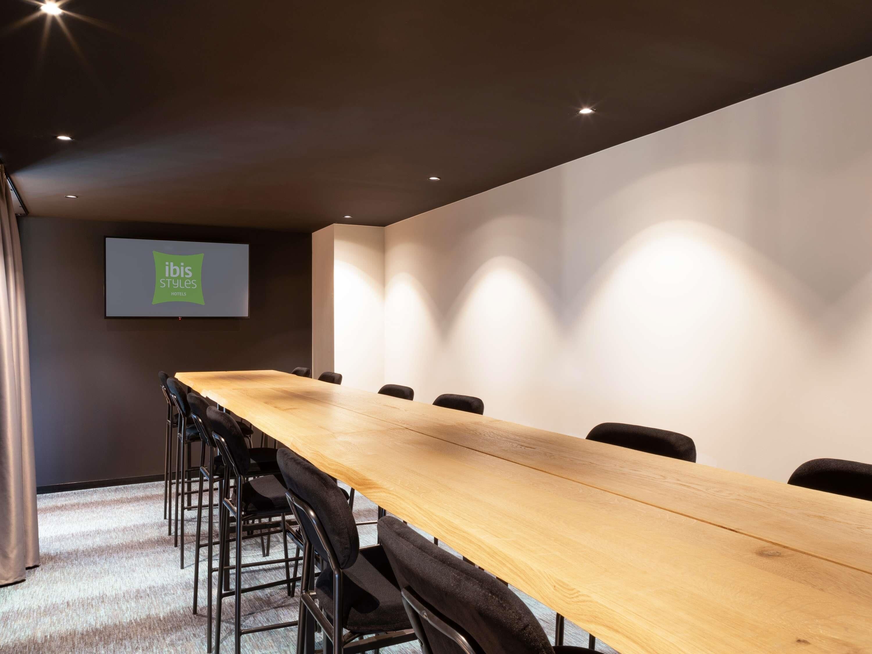 Een vergaderruimte met een lange tafel, stoelen, een projectiescherm en een wandgemonteerd scherm waarop 'ibis Styles hotels' staat.