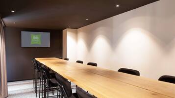 Een vergaderruimte met een lange tafel, stoelen, een projectiescherm en een wandgemonteerd scherm waarop 'ibis Styles hotels' staat.