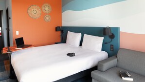 A hotel room with a bed, a desk with a laptop, a grey armchair, and a wall with decorative patterns. - Ibis Styles Miramas - Provence (Miramas)