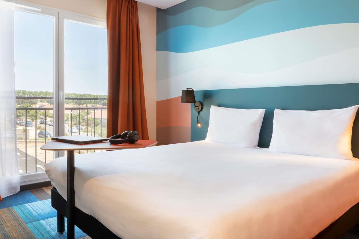 Quarto de hotel com uma cama grande, uma mesinha com telefone, uma luminária na parede e vista para o exterior.