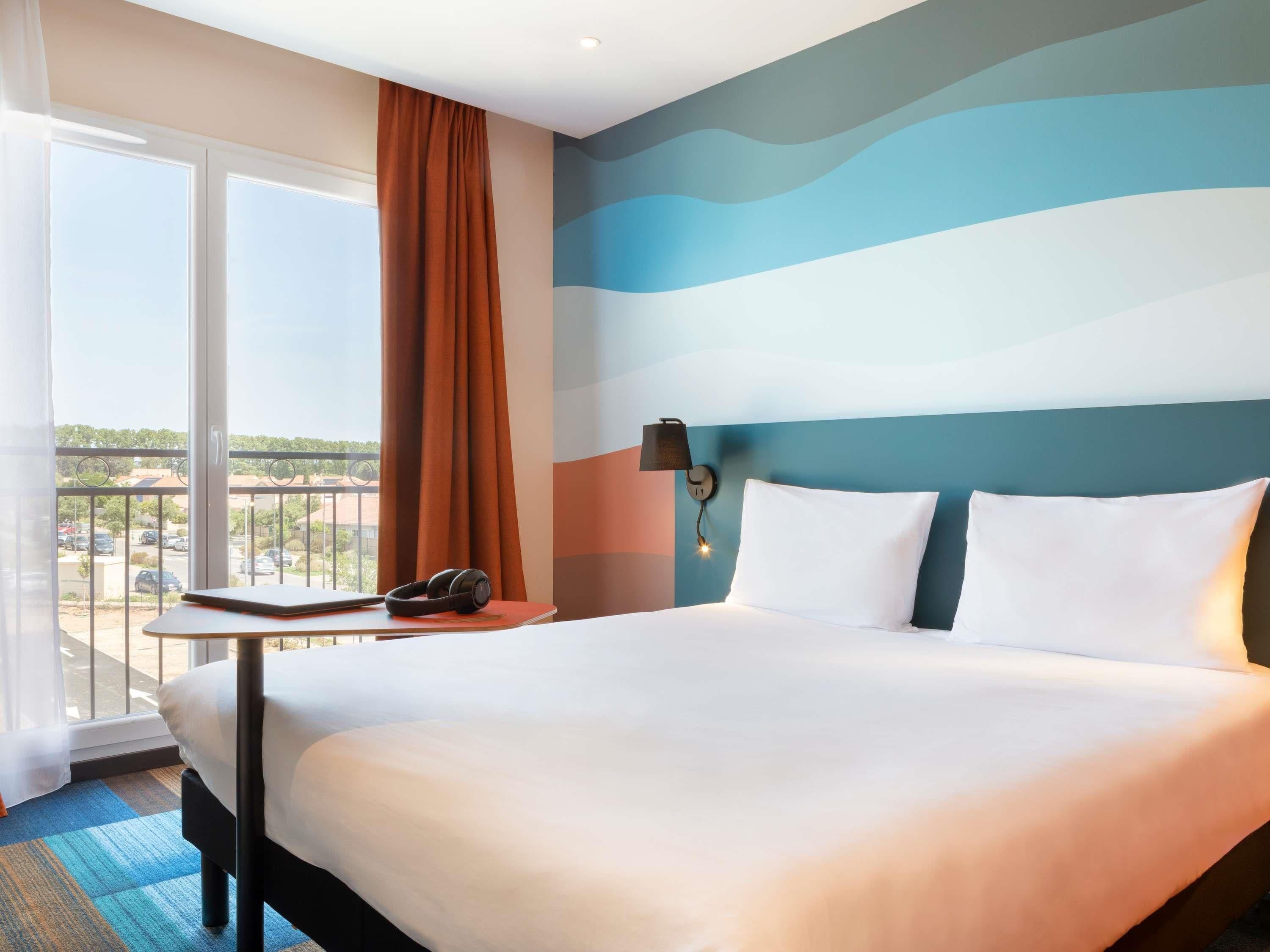 A hotel room with a large bed, a small table with a phone, a wall-mounted lamp, and a view of the outside.