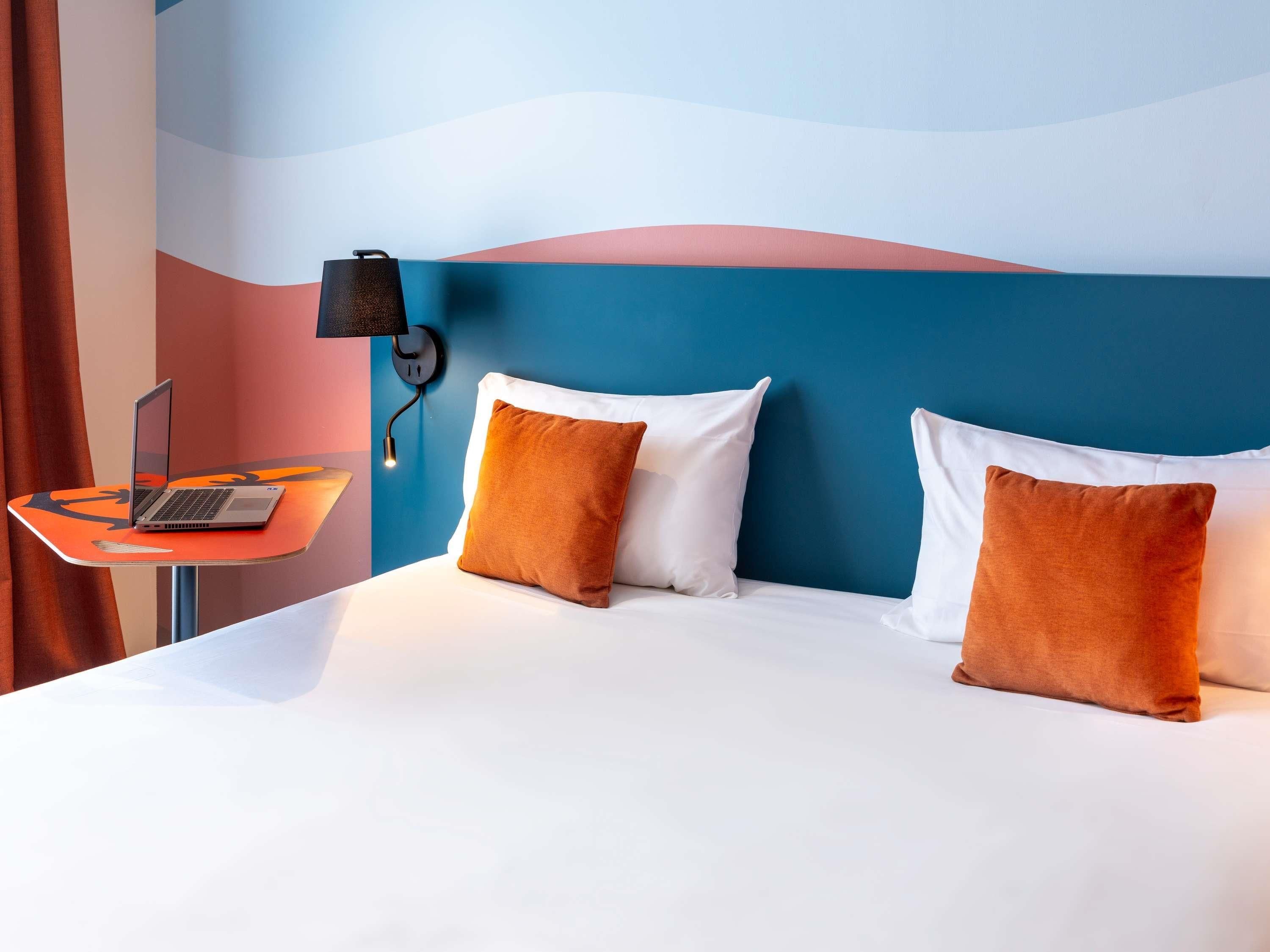 Ein Hotelzimmer mit einem Bett, zwei Kissen, einem Laptop auf einem kleinen Tisch und einer Wandleuchte.