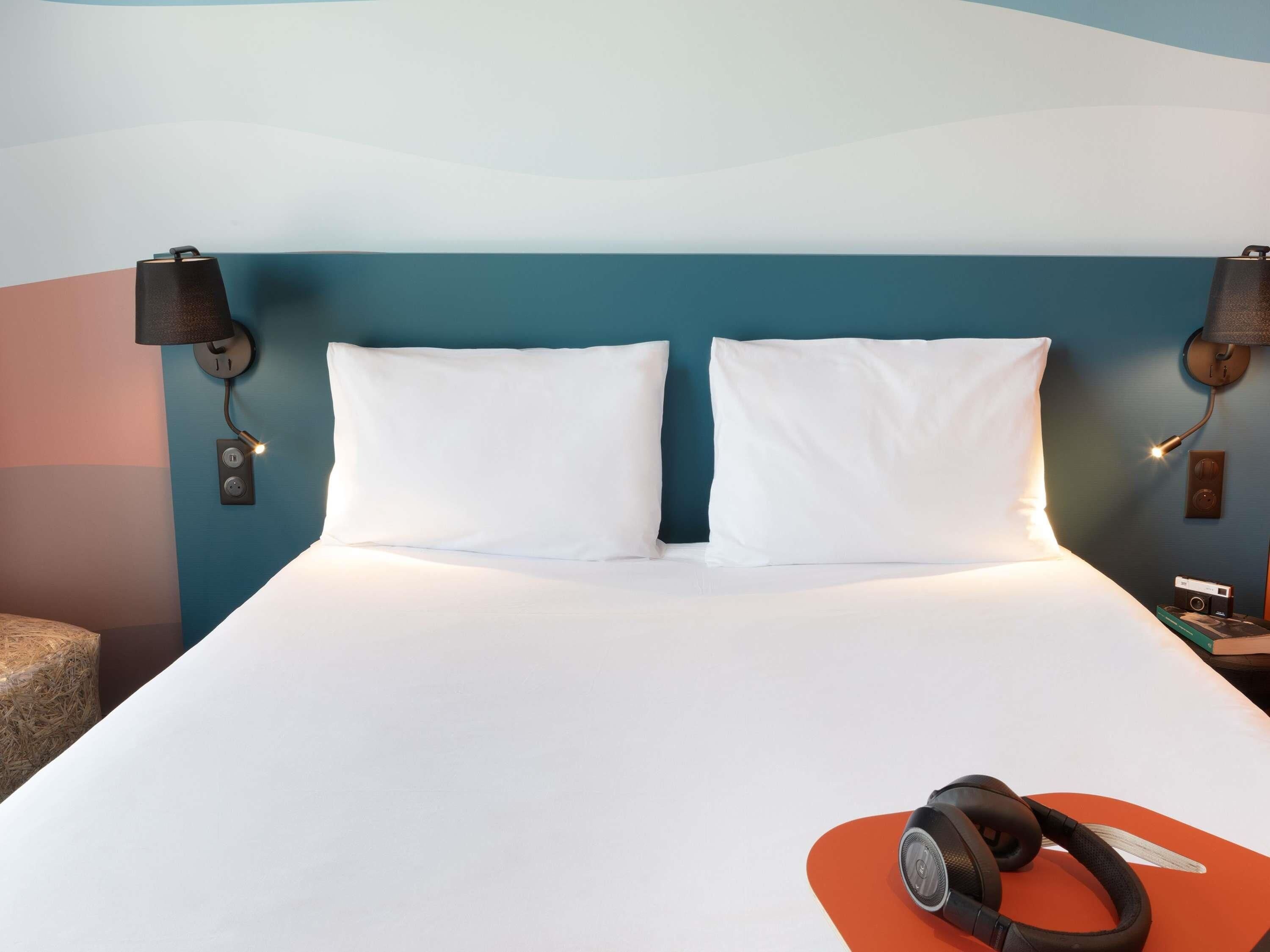 Un lit bien fait, avec des draps et des oreillers blancs, un casque audio posé sur un oreiller orange et une lampe fixée au mur, sur le côté gauche.