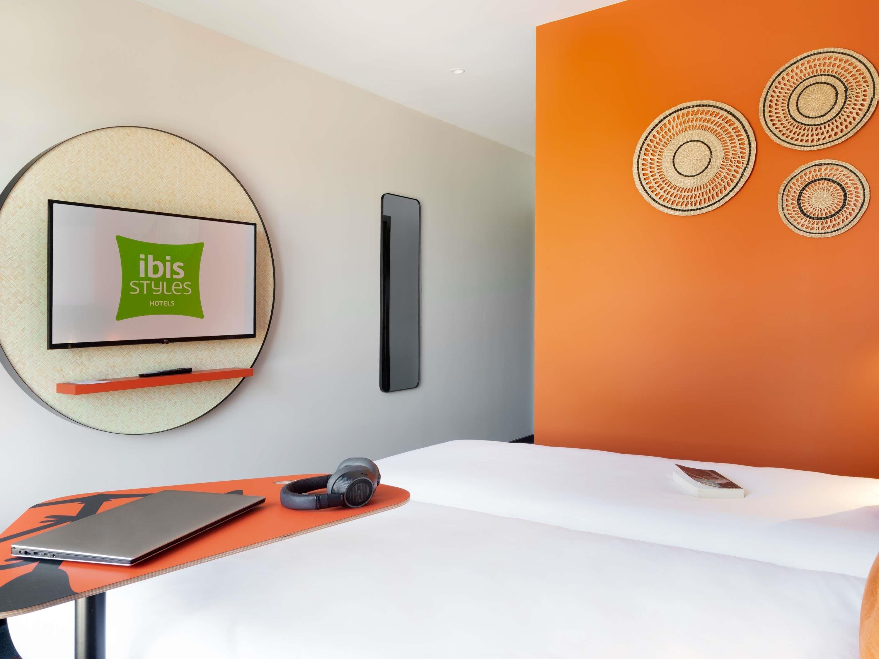 Une chambre d’hôtel moderne avec une horloge murale ronde, un téléviseur à écran plat, une table de chevet avec des écouteurs et un lit recouvert d’une couette blanche.