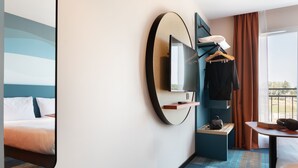A modern hotel room with a large bed, a TV mounted on the wall, a small table with a chair, and a window with curtains. - Ibis Styles Miramas - Provence (Miramas)