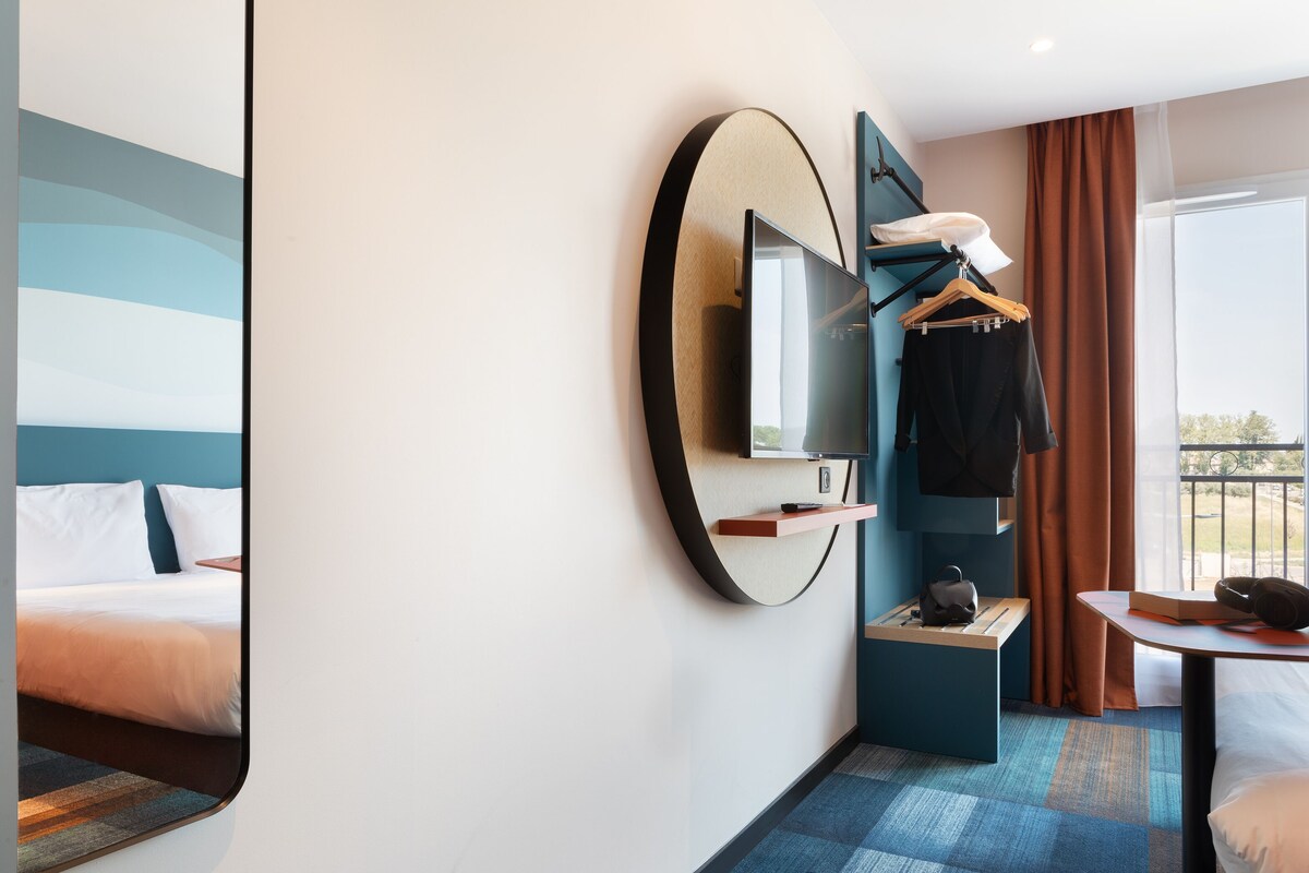 A modern hotel room with a large bed, a TV mounted on the wall, a small table with a chair, and a window with curtains.