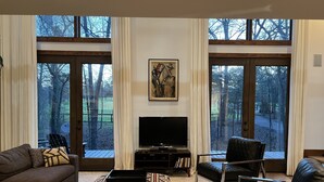 Un salon comprenant un canapé, un pouf et des fauteuils, un téléviseur à écran plat et de grandes fenêtres donnant sur la verdure.