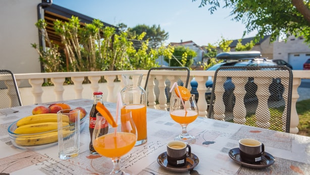 A table set with a glass of orange juice, a bottle of Coca-Cola, a glass of water, a bowl of bananas, and a cup of coffee on a newspaper-covered table. - Apartments Loncar (Vir)