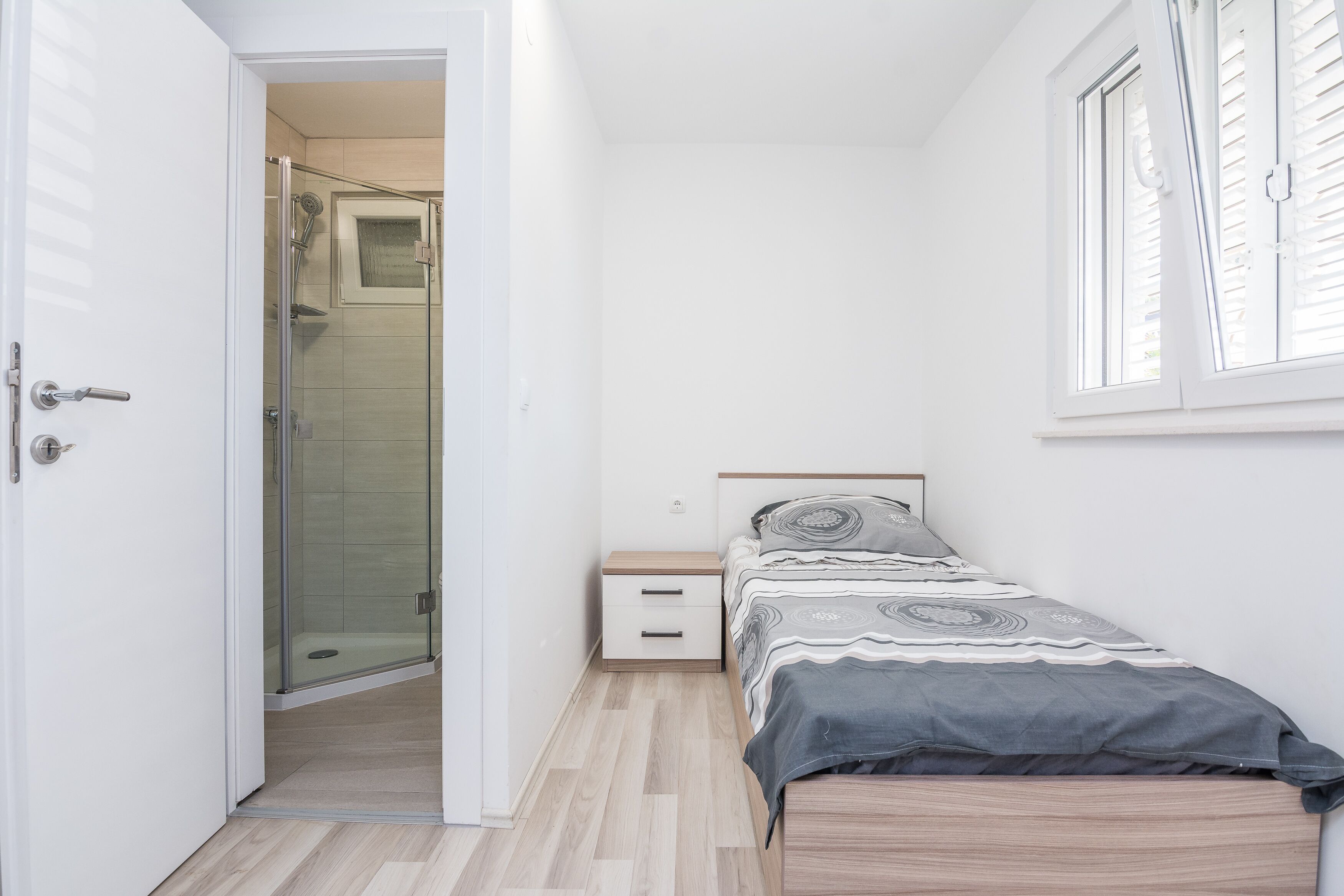 Een slaapkamer met een bed, nachtkastje, jaloezieën en een douchecabine met glazen wanden.