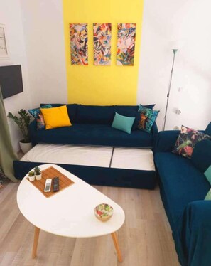Uma sala de estar com um sofá azul, uma mesa de centro branca e três quadros coloridos na parede.