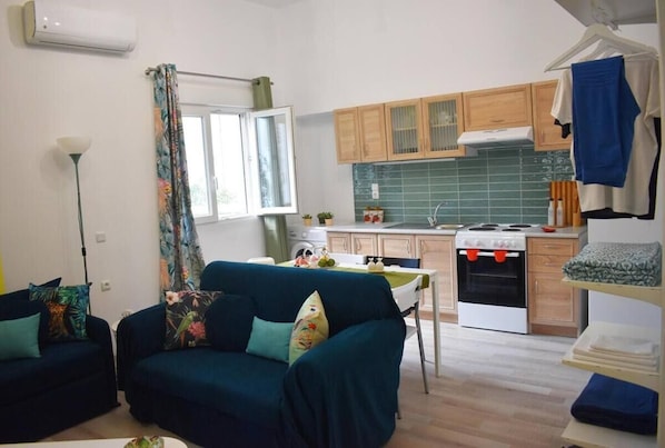 A compact living space with a sofa, a dining table, a kitchenette, and a window with curtains. - Pirgos Colorful Studio, Sleeps 2, Parking Wifi Ac (Archanes-Asterousia)