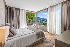 A bedroom with a bed, a bedside table, a lamp, and a view of a balcony with a cityscape and mountains in the background. - RDR234 - Amazing modern villa located minutes from beach (Marbella)