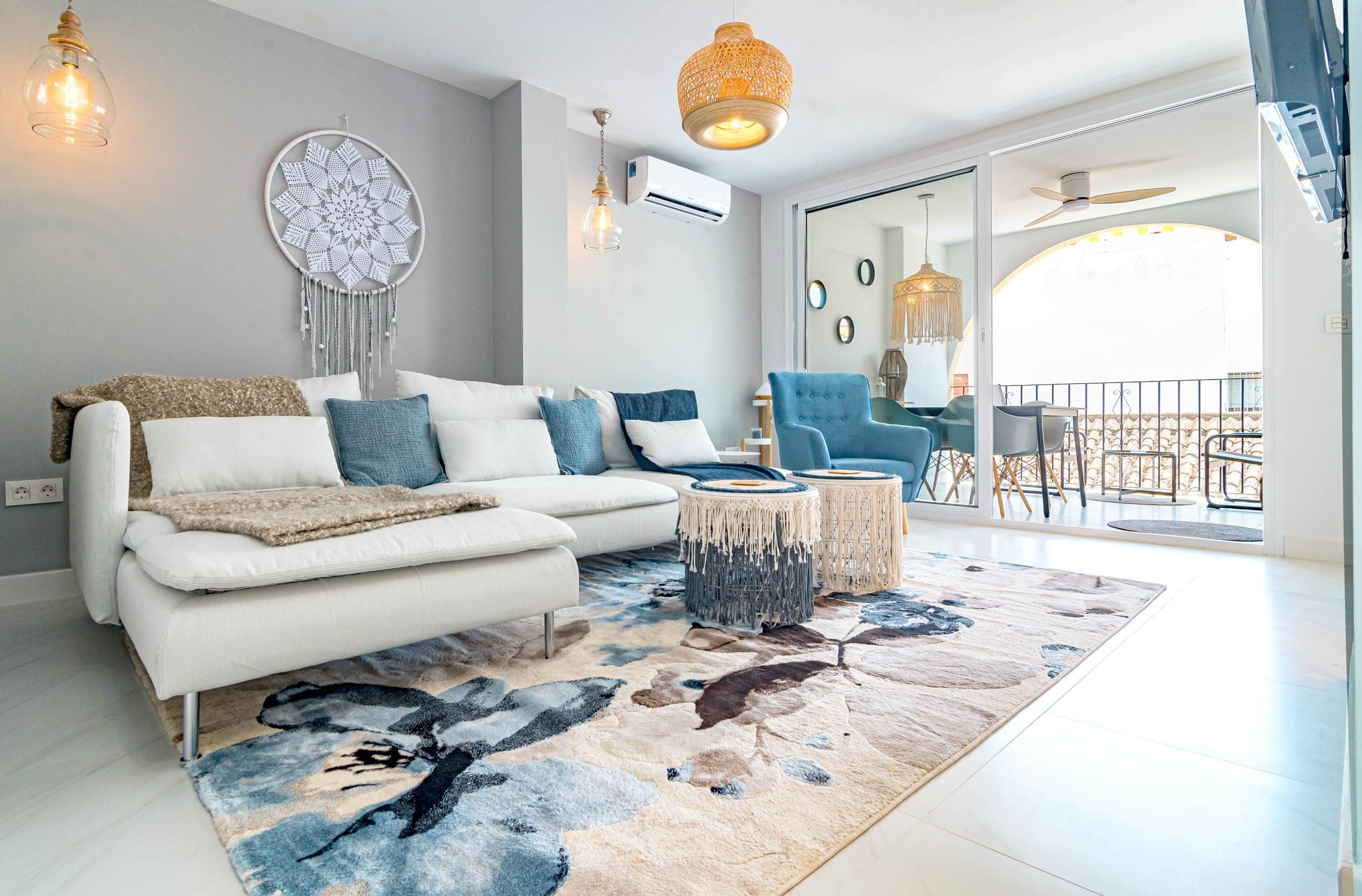 A modern living room with a white sofa, blue cushions, a patterned rug, and a large window offering a view of the outdoors.